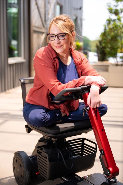 Anna Schmid, a white woman using a mobility scooter, wearing glasses and smiling outside.