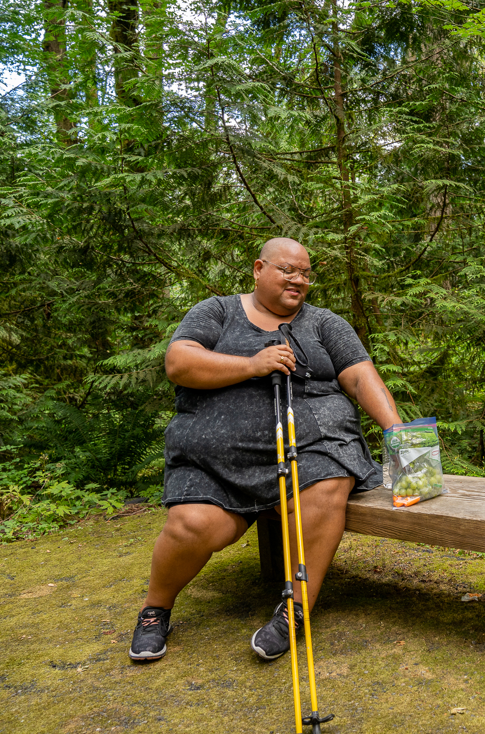 Nonbinary Black woman with walking sticks takes a break on a hiking trail to eat grapes as a snack.